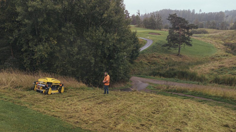 Spider ILD02 -rinneniittokone työskentelee jyrkällä penkereellä puuston reunassa, kuljettaja kauempana tasaisella.